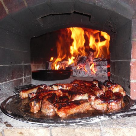 Pork Riblets on a cast iron cooking pan with the wood flame of the Forno Nardona brick oven behind it.