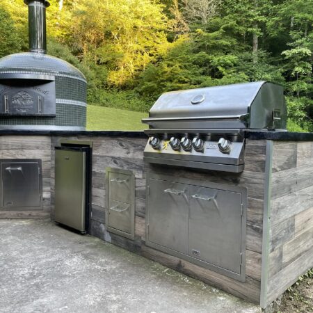 Cabin in the Woods door on a Forno Nardona wood-fired Napoli brick oven in green with white stripes on a rustic wood outdoor kitchen with a black poured concrete countertop featuring a Lion grill, refrigerator and doors/drawers.