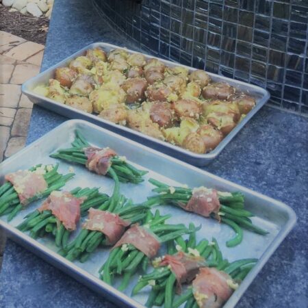 Two trays of food. One with golden garic potatoes with skin and the other with prosciutto wrapped green beans ready to go in the Nardona Napoli oven.