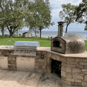 Tampa built outdoor kitchen complete with Lion Grill, Lion accessories and a Forno Nardona Rustico oven with copper tile and a beautiful backdrop of the ocean just beyond the green grass. and trees.
