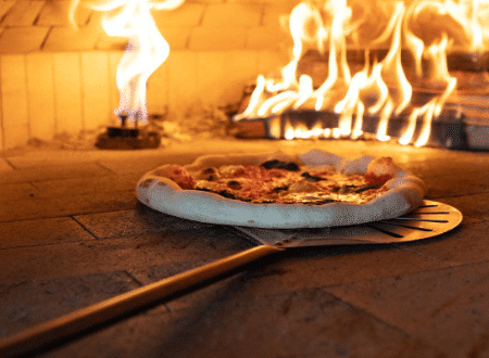 Pizza cooking in a Forno Nardona Commercial Oven with both a wood flame and a gas flame in the background and pizza resting on a metal peel in the foreground.