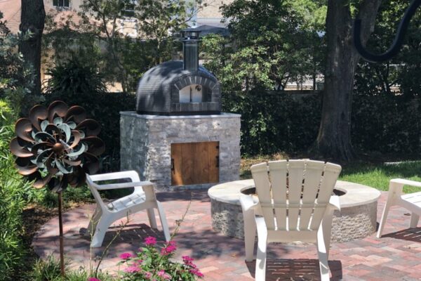 32" Nardona Rustico in grey tile sits atop a custom base also by Forno Nardona with woods in the background and a fire pit with chairs in the foreground.
