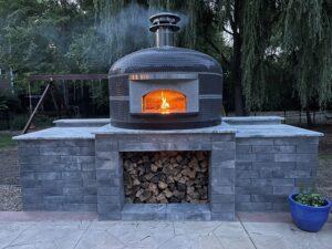 This is one of Forno Nardona's Napoli wood-fired pizza ovens on a customer built stone finished base with wood storage underneath and a flame inside the oven dome.