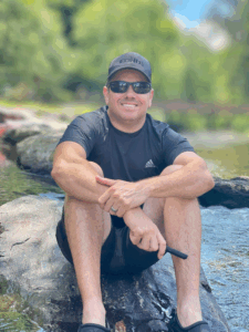 William Hall, owner of Forno Nardona, relaxing on a river rock wearing a baseball cap and sunglasses.